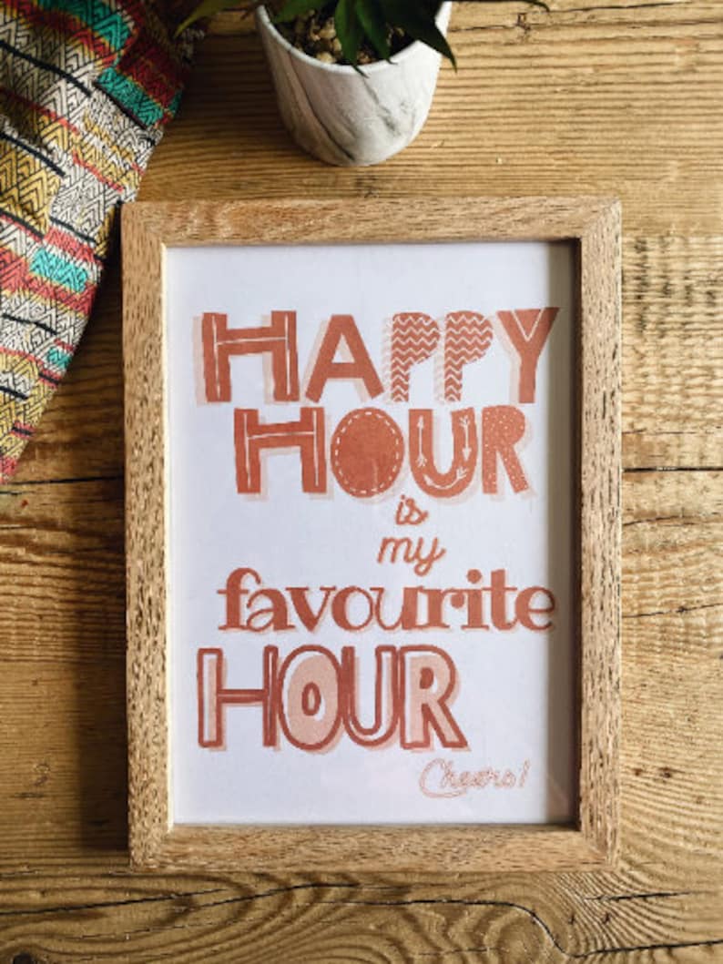 May include: A framed print with the text "Happy Hour is my favourite Hour Cheers!" in a peachy orange color. The print is in a light wood frame.
