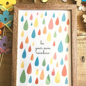 May include: A framed print with a white background and colorful raindrops. The print says "be your own rainbow".