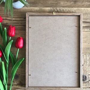 May include: A brown wooden picture frame with a light brown cardboard backing. The frame is sitting on a wooden surface.