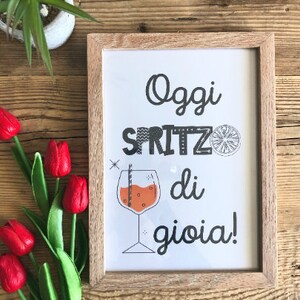 May include: A framed print with the text "Oggi Spritz di gioia!" in black and white with a stylized orange slice and a glass of orange juice with a straw. The frame is made of light brown wood.