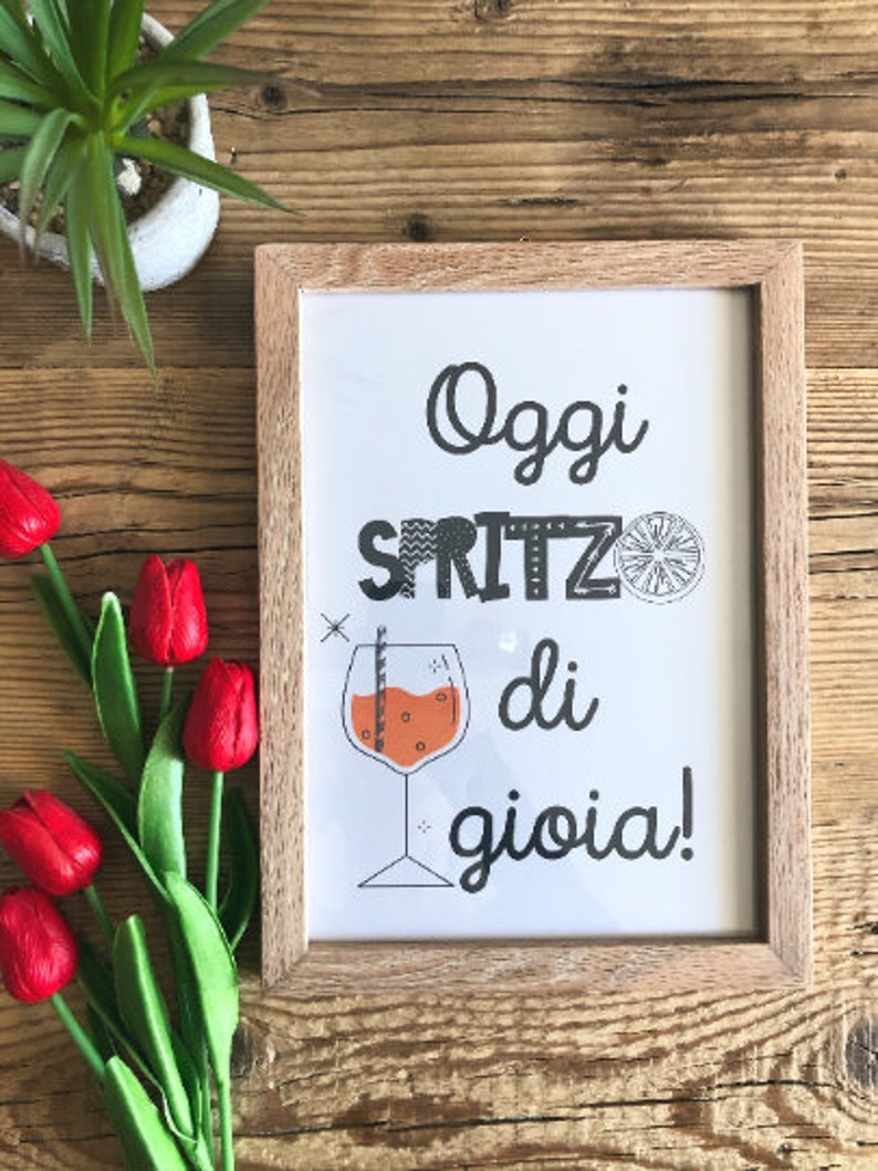 May include: A framed print with the text "Oggi Spritz di gioia!" in black and white with a stylized orange slice and a glass of orange juice with a straw. The frame is made of light brown wood.