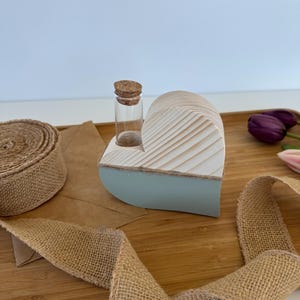 Puede incluir: Un jarrón de madera en forma de corazón con un acabado de pintura azul claro y blanco. Un pequeño vial de vidrio con un tapón de corcho se inserta en la parte superior del jarrón. El jarrón está sentado sobre una superficie de madera con una cinta de arpillera.