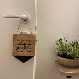 Può includere: Un cartello di legno con la scritta "due gocce di acqua santa e sono pronta" appeso a una porta. Il cartello è marrone con lettere nere.