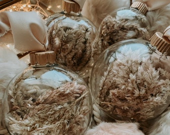May include: Close-up of clear glass Christmas baubles filled with dried beige and pink flowers. Each bauble has a gold-coloured cap and is tied with a cream-coloured ribbon. The baubles are on a bed of fluffy, light-coloured fabric.