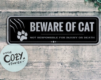 Może przedstawiać: Czarno-srebrny metalowy znak z napisem "Beware of Cat" i słowami "Not responsible for injury or death" poniżej. Znak ma odcisk łapy i ślady pazurów na tle.
