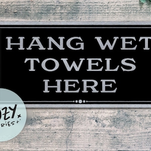 Op de afbeelding: Een rechthoekig zwart en zilveren bord met de woorden "HANG WET TOWELS HERE" in grote, zilveren letters. Het bord heeft een zilveren rand en staat tegen een verweerde, grijze houten achtergrond. Een klein, rond logo staat linksonder.