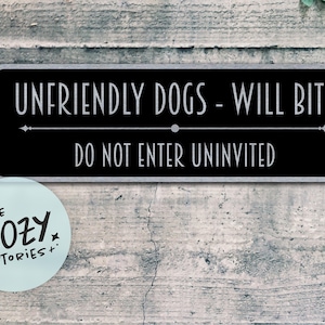 以下が含まれることがあります： 「UNFRIENDLY DOGS - WILL BITE」と「DO NOT ENTER UNINVITED」という白文字が書かれた黒い長方形の看板。左下には「LITTLE COZY STORIES+」という文字が入った水色の丸いステッカーがあります。