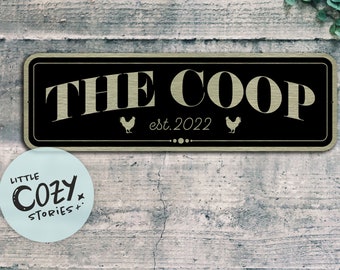 Peut inclure: Une enseigne noire et dorée avec le texte "THE COOP est. 2022" et deux silhouettes de poulets. L'enseigne est fixée à un mur en bois gris.