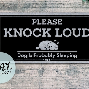 以下が含まれることがあります： シルバーの縁取りと「PLEASE KNOCK LOUD」の文字が入った黒い長方形の看板。その下に、眠っている犬のイラストと「Dog Is Probably Sleeping」の文字があります。左下には丸いロゴがあります。