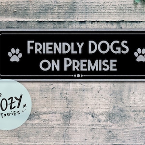 May include: Black rectangular sign with the text "FRIENDLY DOGS ON PREMISE" in white, flanked by paw prints. A small, round sticker with the text "LITTLE COZY STORIES+" is in the lower left corner.