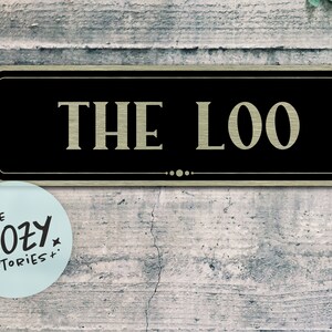 Può includere: Un cartello rettangolare nero e dorato con il testo "THE LOO" in lettere dorate. Il cartello è montato su una parete di legno grigia.