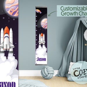 May include: A customizable growth chart featuring a rocket ship launching into space with planets and stars. The chart is printed on a white background with a wood-like frame. The name "Jaxon" is printed in purple at the bottom of the chart.