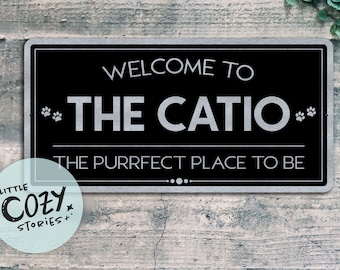 May include: A black and silver metal sign with the text "Welcome to The Catio. The Purrfect Place to Be" and paw prints on either side of the text.