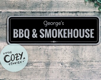 May include: Black and silver sign with the text "George's BBQ & Smokehouse" on a wooden wall. A small sticker with the text "Little Cozy Stories" is below the sign.