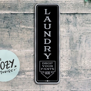 May include: Black and silver metal sign with the word "LAUNDRY" written vertically in silver letters. The sign also says "DROP YOUR PANTS HERE" with an arrow pointing down.