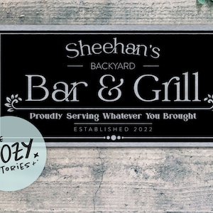 May include: Black metal sign with silver lettering that reads "Sheehan's Backyard Bar & Grill. Proudly Serving Whatever You Brought. Established 2022."