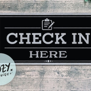 May include: Black rectangular sign with the words "CHECK IN HERE" in white, with a clipboard and pencil icon. The sign has a silver border and is set against a weathered wood background. A small, round logo with the text "LITTLE COZY STORIES+" is in the lower left corner.