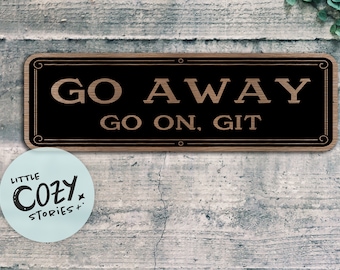 May include: A black wooden sign with a white border and gold lettering that reads "GO AWAY GO ON, GIT". The sign is mounted on a gray wooden wall.
