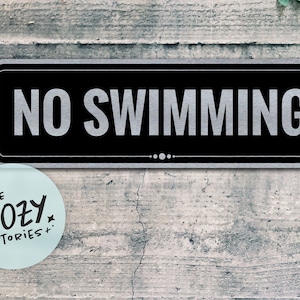 Puede incluir: Un letrero negro y plateado con el texto "NO SWIMMING" en letras blancas. El letrero está montado en una pared de madera.