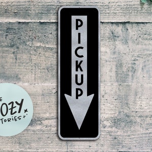 Op de afbeelding: Een zwart en zilveren metalen bord met een witte pijl die naar beneden wijst en het woord "PICKUP" verticaal in witte letters geschreven.