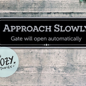 May include: A rectangular black sign with white text that reads "APPROACH SLOWLY" and "Gate will open automatically." A light blue circular sticker with the text "LITTLE COZY STORIES+" is below the sign. The sign is mounted on a textured gray wall.
