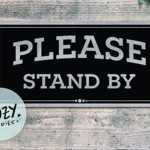 Peut inclure: Panneau rectangulaire noir avec les mots "PLEASE STAND BY" en blanc, soulignés de gris. Le panneau a une bordure grise et est placé sur un fond en bois gris vieilli. Un logo circulaire est dans le coin inférieur gauche.