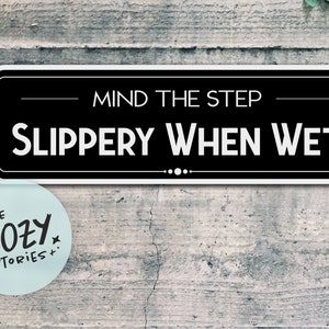May include: Black and white sign with white text that reads "Mind the Step - Slippery When Wet". The sign is mounted on a gray wood surface. A blue sticker with the text "Little Cozy Stories" is in the lower left corner of the image.