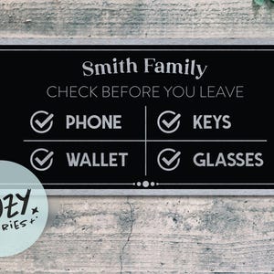 Puede incluir: Un letrero negro y plateado con el texto "Smith Family" y "Check Before You Leave". El letrero tiene casillas de verificación para "Phone", "Keys", "Wallet" y "Glasses". Un logotipo circular de "Little Cozy Stories+" está en la esquina inferior izquierda.