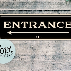 Op de afbeelding: Een zwart-wit bord met het woord "ENTRANCE" in witte letters en een witte pijl die naar links wijst. Het bord is bevestigd aan een grijze houten muur. Een blauwe sticker met de tekst "LITTLE COZY STORIES" bevindt zich onder het bord.