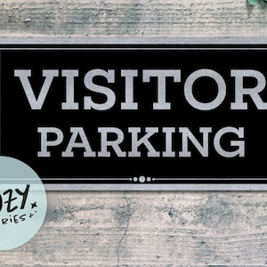 Puede incluir: Un letrero rectangular negro con las palabras "VISITOR PARKING" en letras grandes plateadas. El letrero tiene un borde plateado y está sobre un fondo de madera gris desgastada. Un pequeño logotipo circular con el texto "LITTLE COZY STORIES+" está en la esquina inferior izquierda.