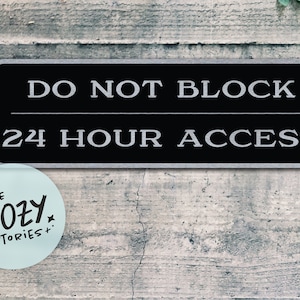 Könnte beinhalten: Ein schwarzes und silbernes Schild mit weißem Text, auf dem "NICHT BLOCKIEREN 24-STUNDEN-ZUGRIFF" steht. Das Schild ist an einer grauen Betonwand angebracht. Unter dem Schild befindet sich ein blauer Aufkleber mit dem Text "LITTLE COZY STORIES".