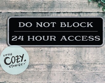 May include: A black and silver sign with white text that reads "DO NOT BLOCK 24 HOUR ACCESS". The sign is mounted on a gray concrete wall. A blue sticker with the text "LITTLE COZY STORIES" is below the sign.
