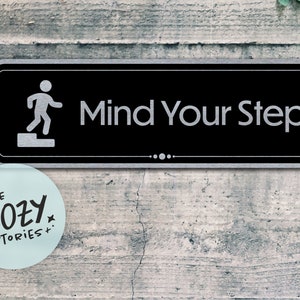 May include: A black and silver sign with the text "Mind Your Step" and a white silhouette of a person walking up stairs.