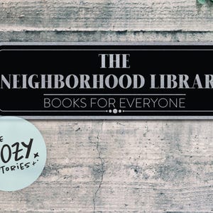 Puede incluir: Un letrero rectangular negro con texto blanco que dice "THE NEIGHBORHOOD LIBRARY BOOKS FOR EVERYONE". Una pegatina circular azul claro debajo dice "LITTLE COZY STORIES+". El letrero está sobre un fondo de madera gris desgastada.