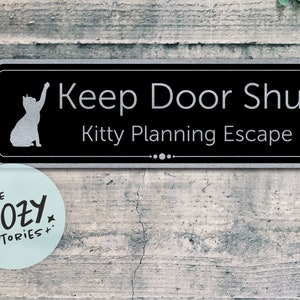 Può includere: Un cartello nero e argento con il testo "Keep Door Shut Kitty Planning Escape" e una silhouette di un gatto con la zampa alzata.