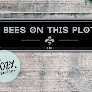 Può includere: Un cartello rettangolare nero con angoli arrotondati che presenta il testo bianco BEES ON THIS PLOT e un'ape grafica centrale. Sotto il testo c'è una linea orizzontale con un piccolo elemento decorativo. Un logo azzurro riporta la scritta LITTLE COZY STORIES.