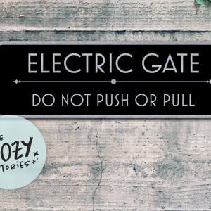 May include: A rectangular black sign with the words "ELECTRIC GATE" in white, above "DO NOT PUSH OR PULL". The sign has a silver border and is mounted on a textured gray wall. A light blue sticker with the text "LITTLE COZY STORIES+" is in the lower left corner.