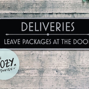 Könnte beinhalten: Ein schwarzes rechteckiges Schild mit weißem Text: „DELIVERIES LEAVE PACKAGES AT THE DOOR“. Das Schild hat einen silbernen Rand und ist an einer verwitterten, grauen Holzwand angebracht. Ein runder Aufkleber mit dem Text „LITTLE COZY STORIES+“ befindet sich links unten.