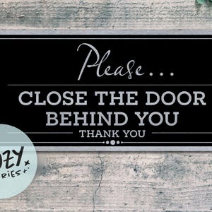 Op de afbeelding: Een zwart rechthoekig bord met een zilveren rand en witte tekst: "Please... CLOSE THE DOOR BEHIND YOU THANK YOU." Een klein, rond, lichtblauw logo met de tekst "LITTLE COZY STORIES+" staat linksonder.