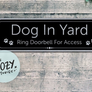 May include: A black and silver sign with the text "Dog In Yard" and "Ring Doorbell For Access". Two paw prints are on either side of the text. The sign is mounted on a wooden surface.