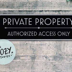 Può includere: Un cartello rettangolare nero con le scritte bianche "PRIVATE PROPERTY" e "AUTHORIZED ACCESS ONLY". Un piccolo adesivo circolare azzurro con la scritta "LITTLE COZY STORIES+" si trova in basso a sinistra. Il cartello è montato su un muro grigio usurato.