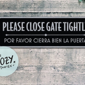 May include: A rectangular black sign with white text that reads "PLEASE CLOSE GATE TIGHTLY" above "POR FAVOR CIERRA BIEN LA PUERTA". A light blue circular sticker with the text "LITTLE COZY STORIES+" is in the lower left corner.