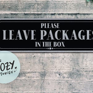 Op de afbeelding: Een rechthoekig zwart en zilveren bord met de tekst "PLEASE LEAVE PACKAGES IN THE BOX". Een ronde sticker met de tekst "LITTLE COZY STORIES+" staat onder het bord. Het bord is bevestigd op een verweerde, grijze houten muur.