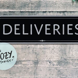 May include: A black rectangular sign with the word "DELIVERIES" in white, set against a textured gray background. A small, light blue circular sticker with the text "LITTLE COZY STORIES+" is in the lower left corner.