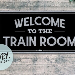 Könnte beinhalten: Schwarzes rechteckiges Schild mit silbernem Text "WELCOME TO THE TRAIN ROOM". Das Schild hat einen silbernen Rahmen und ist an einer verwitterten grauen Holzwand angebracht. Ein kleines, rundes Logo mit dem Text "LITTLE COZY STORIES+" befindet sich in der unteren linken Ecke.