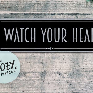 以下が含まれることがあります： 黒い長方形の看板に白い文字で「WATCH YOUR HEAD」と書かれており、風化した灰色の壁に取り付けられています。テキストの下には3つの小さな白い点があります。「LITTLE COZY STORIES+」というテキストの丸いステッカーが左下にあります。