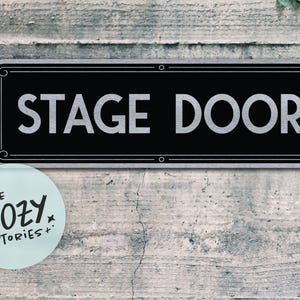 May include: A black rectangular sign with the words "STAGE DOOR" in silver lettering. The sign has a silver border and is mounted on a weathered, gray-toned wall. A light blue circular sticker with the text "LITTLE COZY STORIES+" is in the lower left corner.