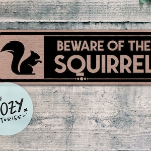 May include: A wooden sign with a black background and white text that reads "Beware of the Squirrel". A silhouette of a squirrel is on the left side of the sign.