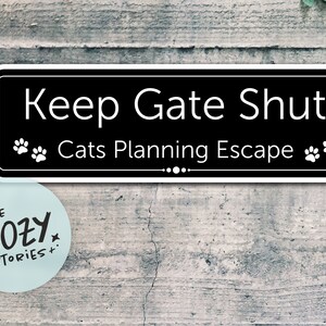 May include: Black and white sign with white text that reads "Keep Gate Shut" and "Cats Planning Escape". The sign has two paw prints on either side of the text. The sign is on a grey wooden background.