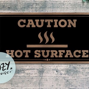 Può includere: Un cartello rettangolare in legno con sfondo nero e le parole "CAUTION HOT SURFACE" in un carattere marrone chiaro e rustico. Il cartello include una grafica di tre linee di vapore sopra una linea orizzontale.